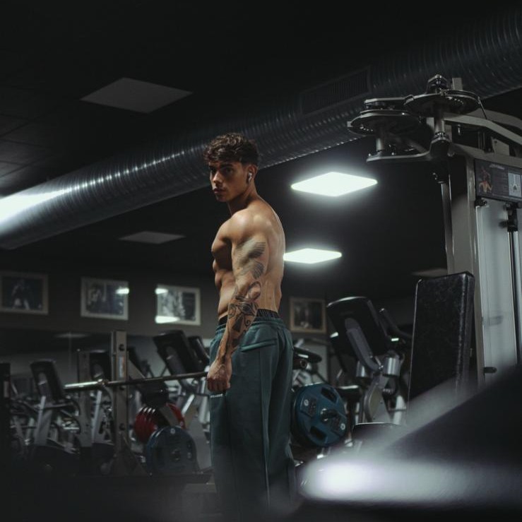 Francesco Sisko Consales in posa laterale in palestra, pantaloni verde petrol, muscoli e tatuaggi in evidenza